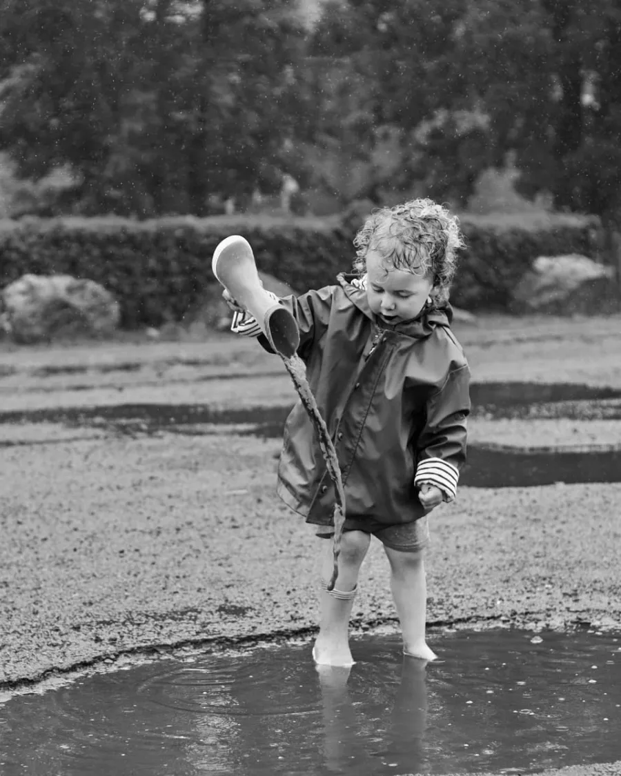 Jeune enfant en ciré vidant sa botte de pluie dans une flaque, en noir et blanc