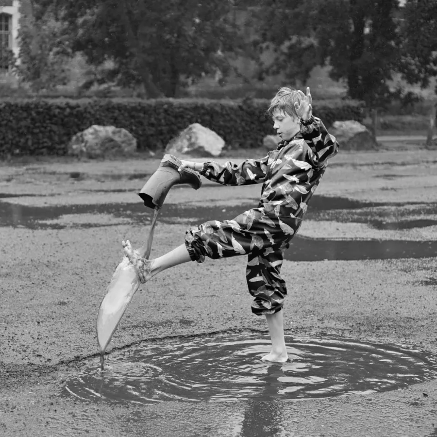 Enfant vidant l'eau de sa botte au-dessus d'une flaque, en noir et blanc
