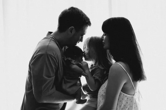 Parents, grande sœur et nouveau-né réunis en contre-jour, en noir et blanc