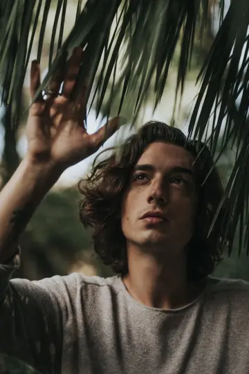 Jeune homme aux cheveux longs ondulés en tee-shirt gris, la main levée vers des feuilles de palmier