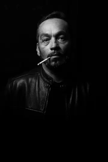 Homme barbu en blouson de cuir, une cigarette au coin des lèvres, sur fond noir, en noir et blanc