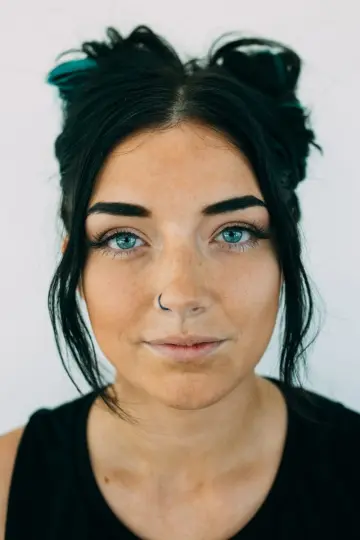 Jeune femme aux yeux bleus, cheveux bruns en deux chignons à mèche turquoise, piercing au nez, sur fond clair