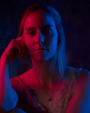 Femme blonde éclairée de lumières rouge et bleue, un doigt posé sur la tempe, sur fond sombre