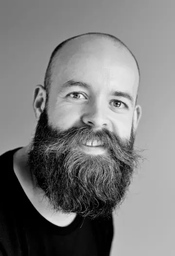 Homme chauve à la longue barbe fournie, souriant, en tee-shirt noir sur fond clair, en noir et blanc