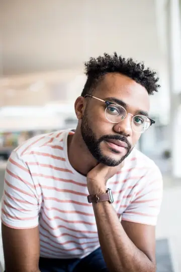 Homme barbu à lunettes rondes en tee-shirt rayé, le menton posé sur la main, dans un intérieur lumineux